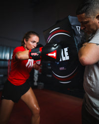 variant:lifestyle::TITLE Boxing Gel Ripple Strike Shield being held by boxing coach with boxer punching shield