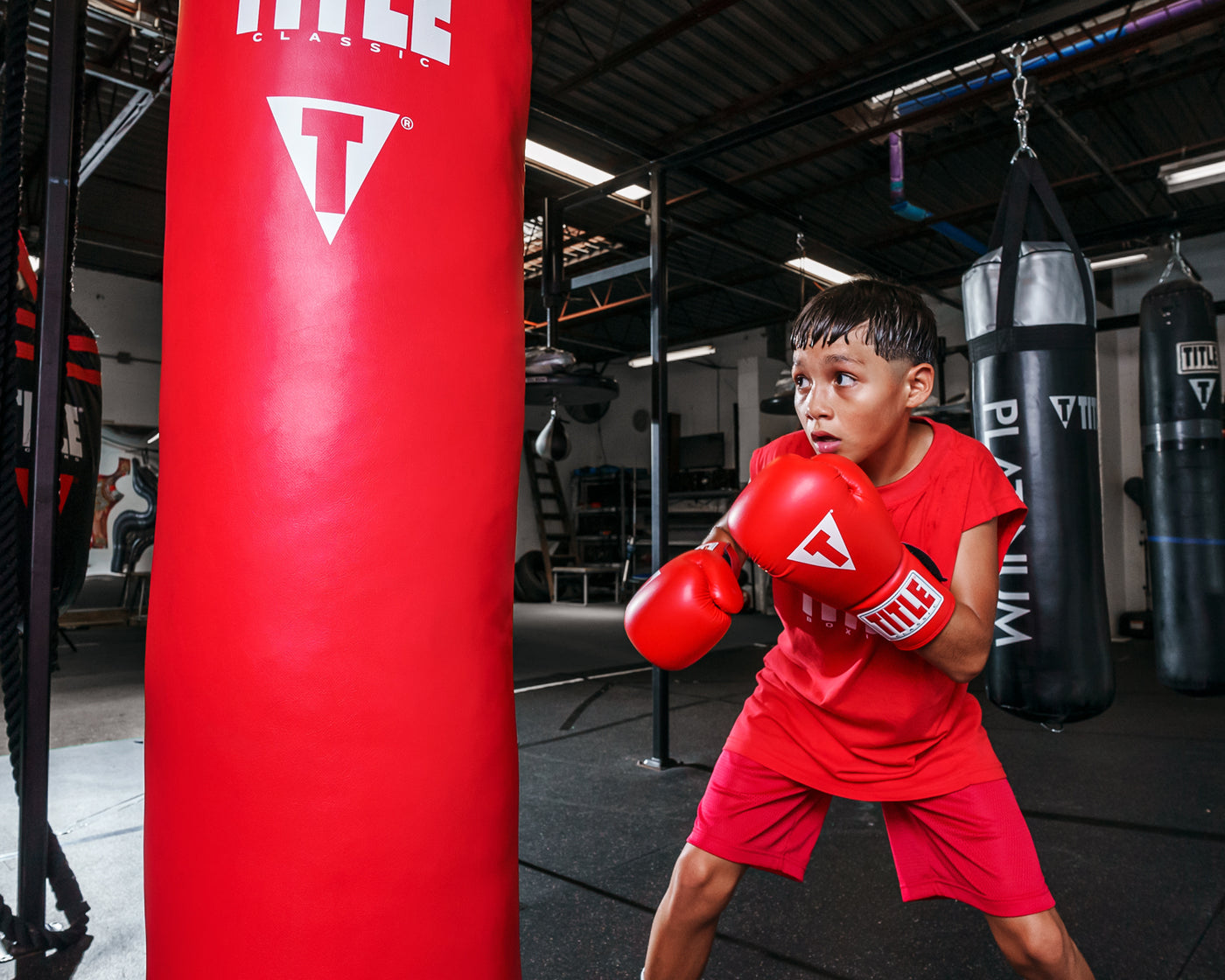 variant:red::TITLE Classic Super Bag Gloves 2.0 kid wearing all red hitting a red bag