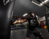 variant:gold/black::TITLE Boxing Infused Foam Interrogate Training Gloves 2.0 guy wearing boxing gloves punching heavy bag