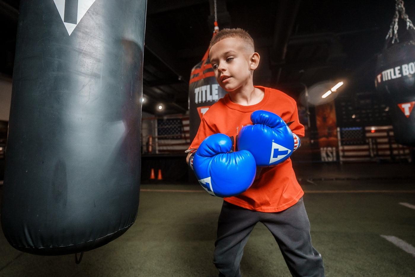 variant:blue/black/white::TITLE Classic Kid & Youth Boxing Gloves 2.0 kid wearing boxing gloves in gym with heavy bags