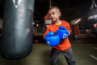 variant:blue/black/white::TITLE Classic Kid & Youth Boxing Gloves 2.0 kid wearing boxing gloves in gym with heavy bags
