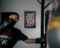 variant:lifestyle::TITLE Boxing Train Fight Win Repeat Banner hanging on a wall behind a boxer training on a heavy bag